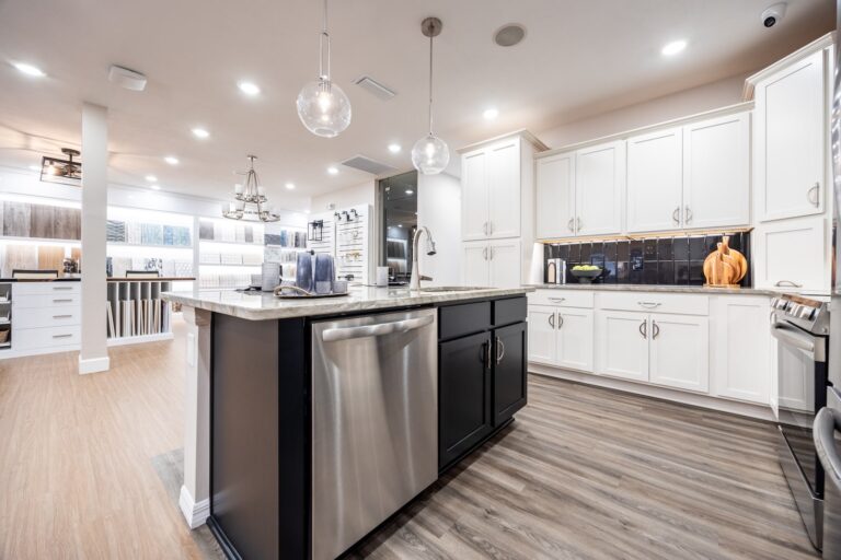 Modern kitchen with white cabinets, a black island featuring a built-in dishwasher, pendant lights, stainless steel appliances, wood flooring, and decorative items including a pumpkin on the counter.