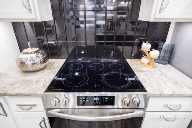 Modern kitchen with a black electric stovetop, stainless steel oven, marble countertop, reflective black tile backsplash, and decorative items including a silver vase and a tray with jars.