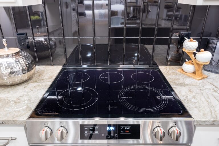 A modern electric stovetop with four burners and a digital display sits on a marble countertop, with black tile backsplash and kitchen containers on either side.