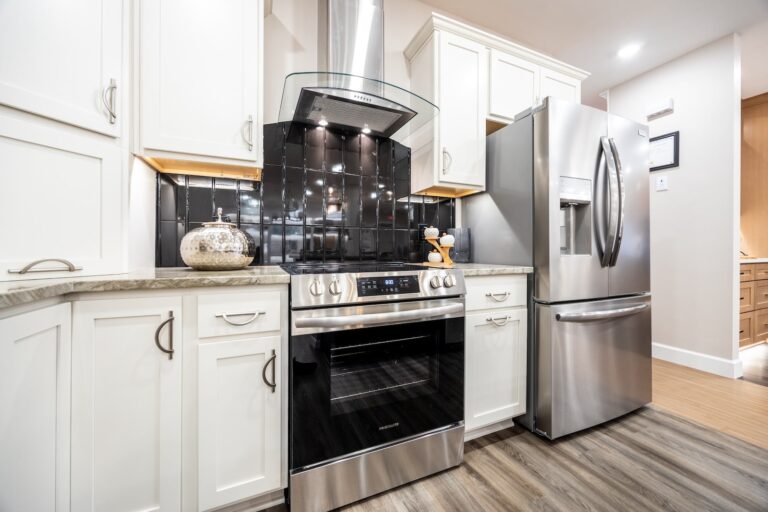 Modern kitchen with white cabinets, stainless steel oven and range hood, black tile backsplash, refrigerator, decorative jar, and wooden utensils on a light-colored countertop. Wood flooring completes the space.