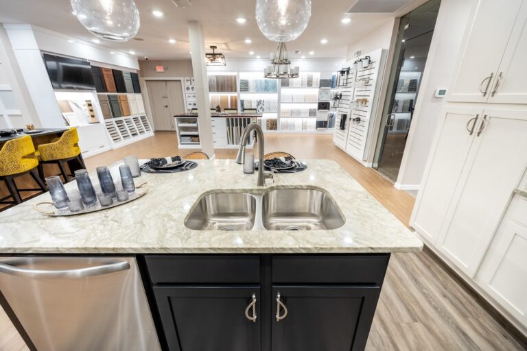 Modern showroom featuring a kitchen display with a marble island, double sink, and black cabinets. Background shows wall and flooring samples, yellow stools, and contemporary lighting.