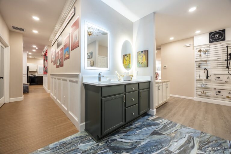 Modern showroom with bright lighting, showcasing a vanity with a round mirror, dark cabinets, and marble-patterned rug. Walls display framed art, and a nearby section features hardware fixtures on white display boards.