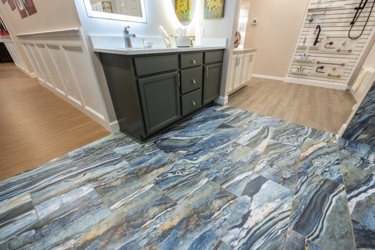 A modern bathroom features blue and gray marble tile flooring, a dark vanity with a white countertop, and a well-lit mirror. The adjacent area has wood flooring and wall displays of faucets and shower fixtures.