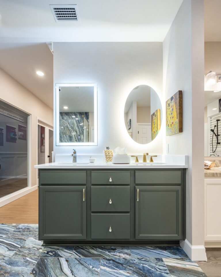 Modern bathroom with dark green vanity, white countertop, two mirrors (one rectangular with lights, one round), gold fixtures, abstract artwork on the wall, and a blue marble-patterned floor.