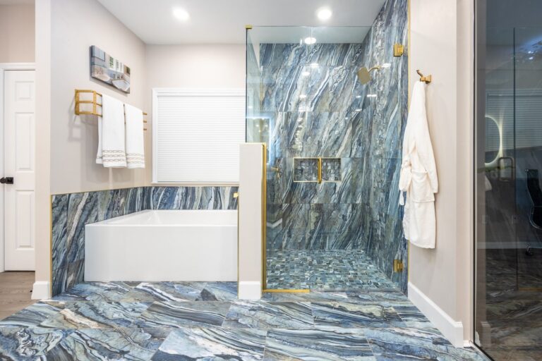 Modern bathroom with a large soaking tub and a glass-enclosed shower featuring striking blue and gray marble tile, gold fixtures, towel racks, and a white bathrobe hanging on the wall.