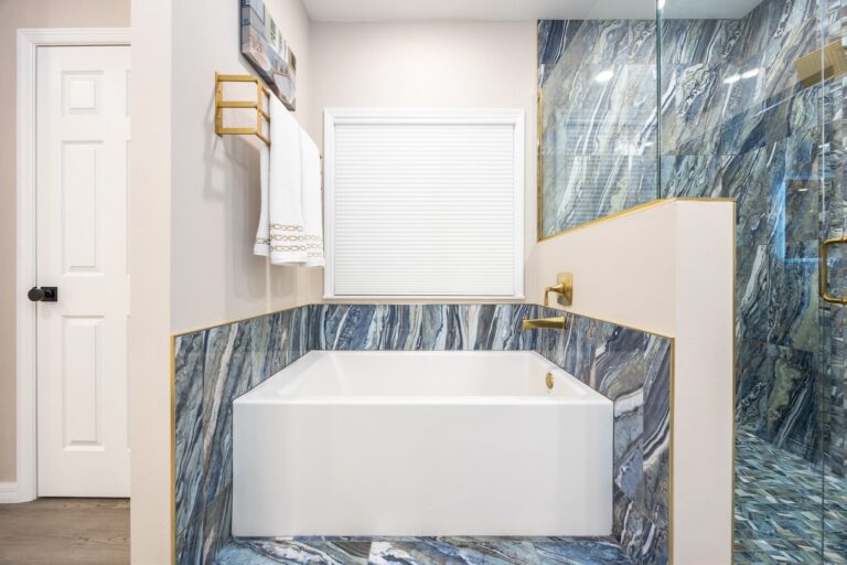 Modern bathroom with a white bathtub set in blue marble tile, gold fixtures, and a glass shower enclosure. White towels hang on a gold rack and a window with blinds is above the tub.