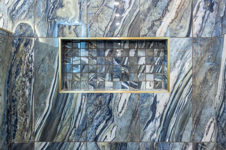 A shower wall with blue and gray marble-like tiles, featuring a recessed niche lined with small square mosaic tiles in matching blue and gray tones.