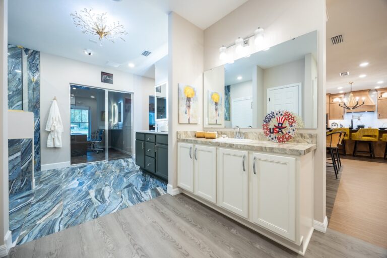 Modern bathroom with white cabinets, marble counters, large wall mirror, blue marble-patterned floor, glass shower, robe hanging on the wall, and a view into a kitchen with barstools and pendant lights in the background.
