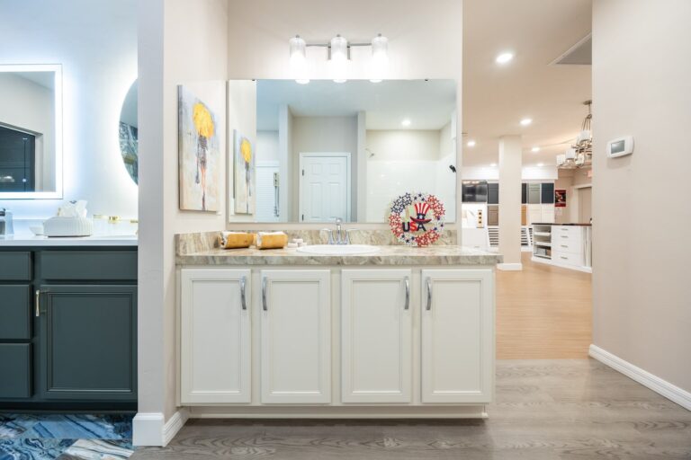 Modern bathroom with a beige and marble vanity, large mirror, and three light fixtures above. The counter has towels and a decorative USA-themed centerpiece. Part of an open kitchen and living area is visible in the background.