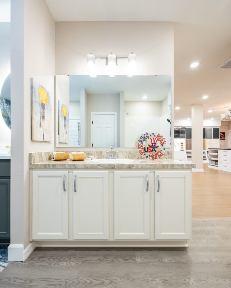 A modern bathroom with a large mirror, three-light fixture, white cabinets, marble countertop, two rolled towels, and a decorative USA sign; neutral walls and wood flooring complete the space.
