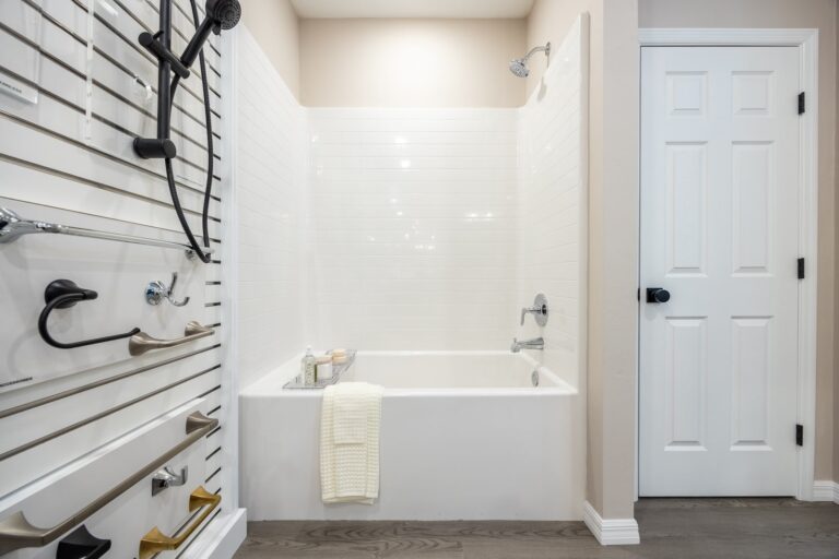 A modern bathroom with a white bathtub, white tiled walls, a hand towel draped over the tub’s edge, and various metal grab bars displayed on a wall. A white door is on the right. The floor is light wood.