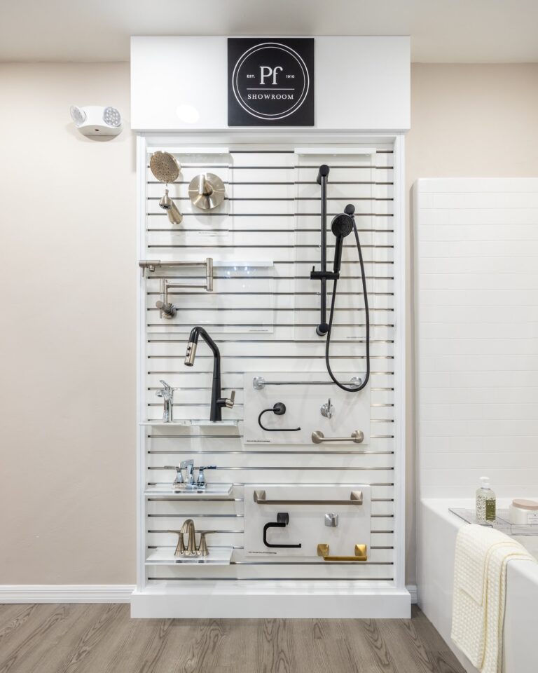 A showroom display wall featuring various modern bathroom and kitchen fixtures, including faucets, showerheads, and handles in different finishes, mounted on white slats. A white bathtub with towels is visible on the right.