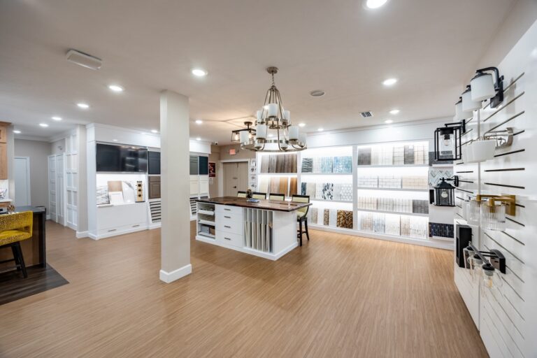 A modern showroom with wood flooring, white walls, display boards showcasing tiles, a central island table, a chandelier, and wall-mounted light fixtures. The space is well-lit and organized.