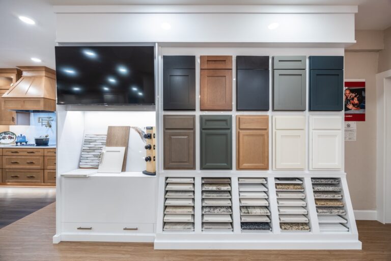 A display wall in a showroom featuring various cabinet door samples in different colors and finishes, with granite and marble countertop samples arranged below and a large TV mounted on the upper left.