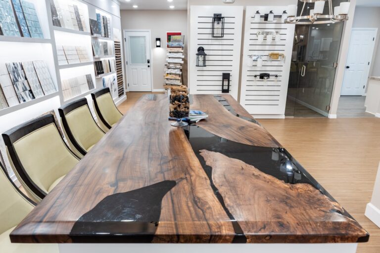 A modern showroom with a large wooden conference table featuring black resin inlays, surrounded by chairs. Display walls show various tile samples and design materials, with bright lighting and wood flooring.
