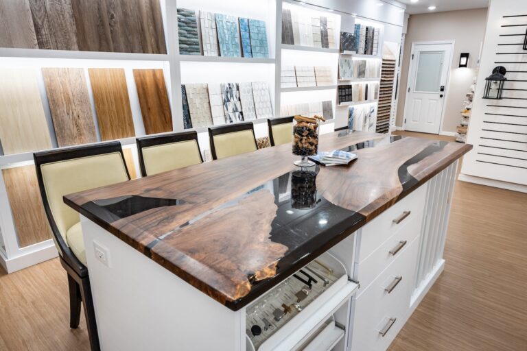 A modern showroom features a wooden island table with a polished live-edge top, surrounded by chairs. Walls display various tile and wood samples, and shelves hold finish options. A jar of cookies sits on the table.