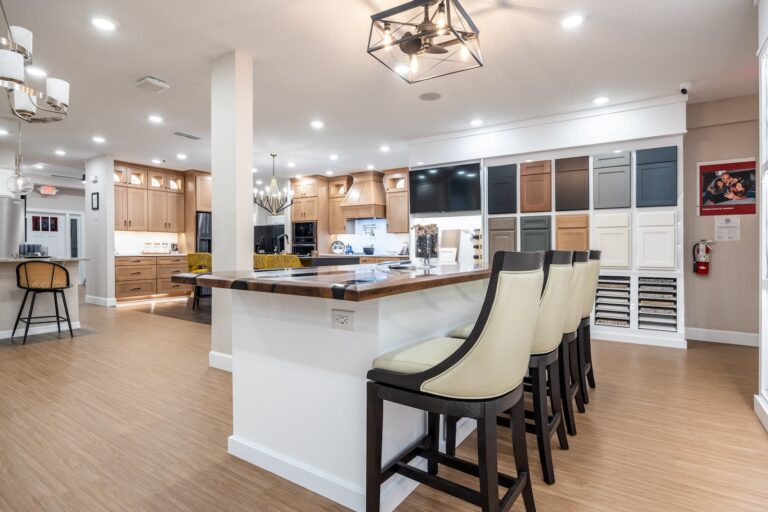 A modern showroom displays kitchen cabinets, countertop samples, and various finishes. A large island with cushioned chairs is centered, surrounded by wood flooring, pendant lights, and wall displays of cabinet doors and colors.