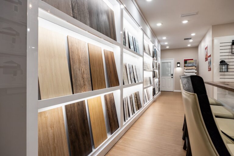 A showroom interior displays various wood and laminate flooring samples mounted on a well-lit wall. The space has light-colored floors, modern lighting, and high-back chairs near a counter.
