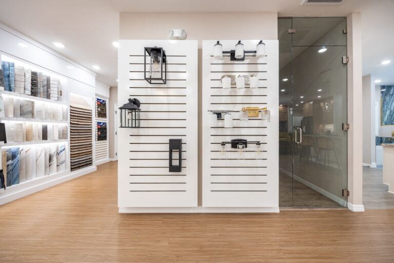 A modern showroom with wood flooring, featuring wall displays of lighting fixtures and samples, and a glass-walled office on the right.