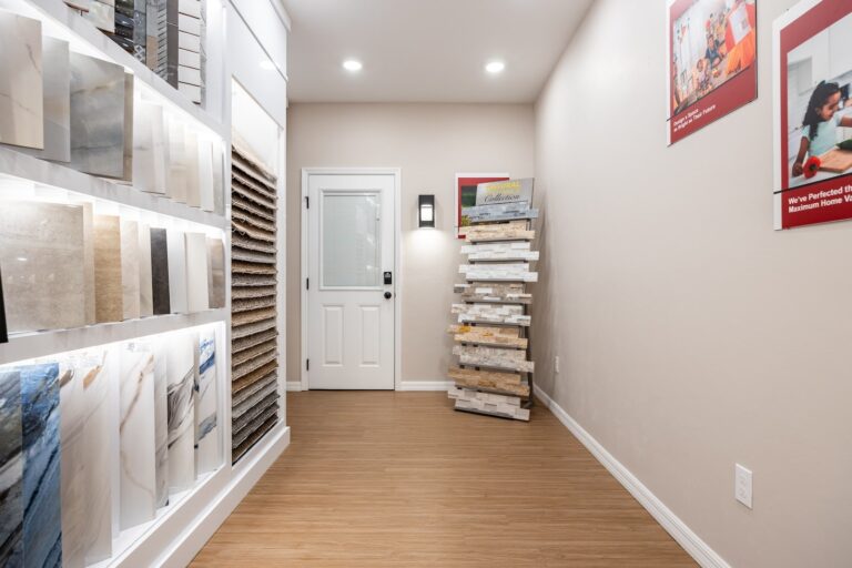 A showroom with wood flooring, walls lined with samples of tiles and countertop materials, a white door at the end, and bright overhead lighting. Red-framed posters hang on the wall.
