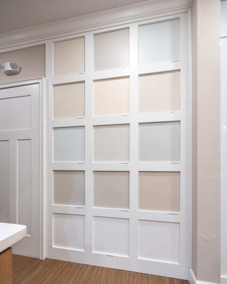A wall display with sixteen square panels framed in white, each panel painted a different neutral shade ranging from beige to light gray, next to a white door and wood flooring.