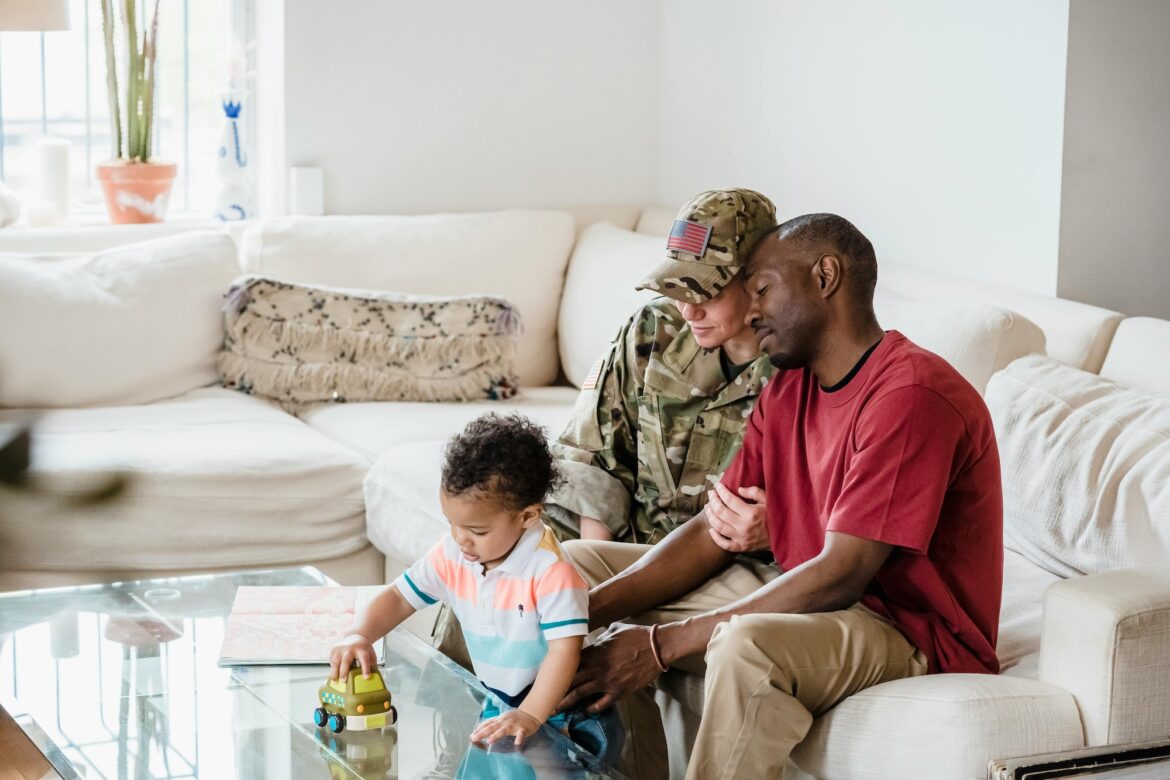 A family sits together in a living room. An adult in military uniform and another in a red shirt sit on a couch, affectionately watching a young child play with a toy on a glass coffee table.
