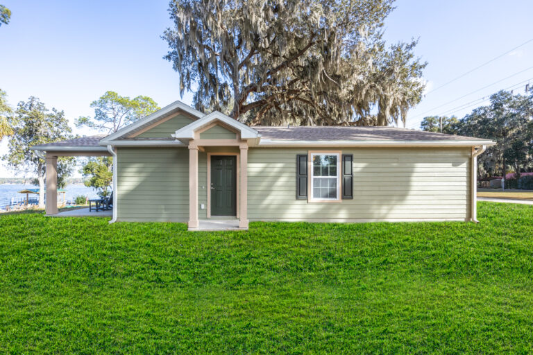 Single-story light green house with a small covered porch, black front door, white-trimmed window, and green lawn; large tree with Spanish moss in the background.