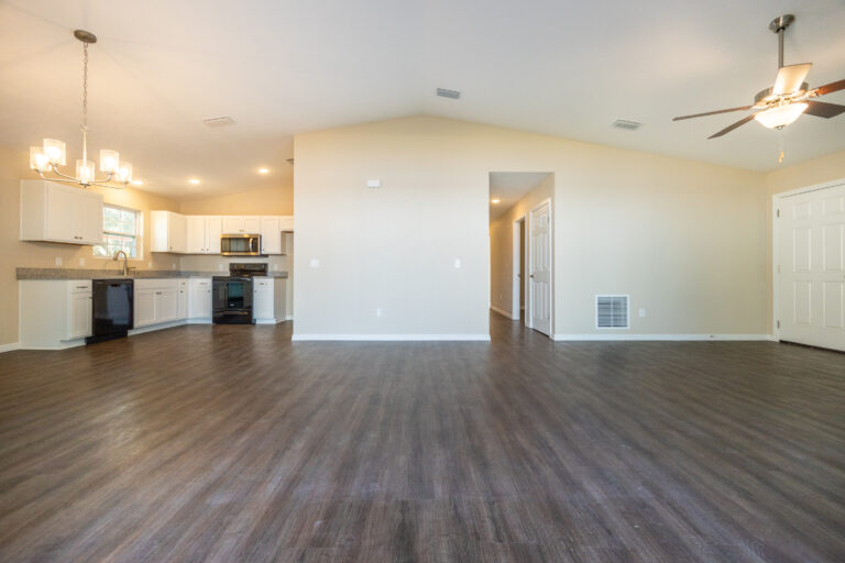 Open concept living area with dark wood floors, beige walls, and a vaulted ceiling. The kitchen features white cabinets, stainless steel appliances, and pendant lighting. A ceiling fan is on the right.