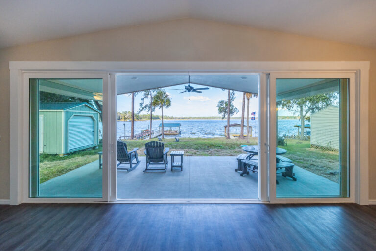 View through large glass doors of a covered patio with Adirondack chairs, overlooking a lake, palm trees, and a shed, on a clear day.
