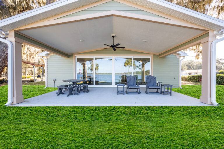 A covered patio with outdoor seating, including Adirondack chairs and picnic tables, overlooks a grassy lawn and a lake, with large glass doors leading into a light green house.