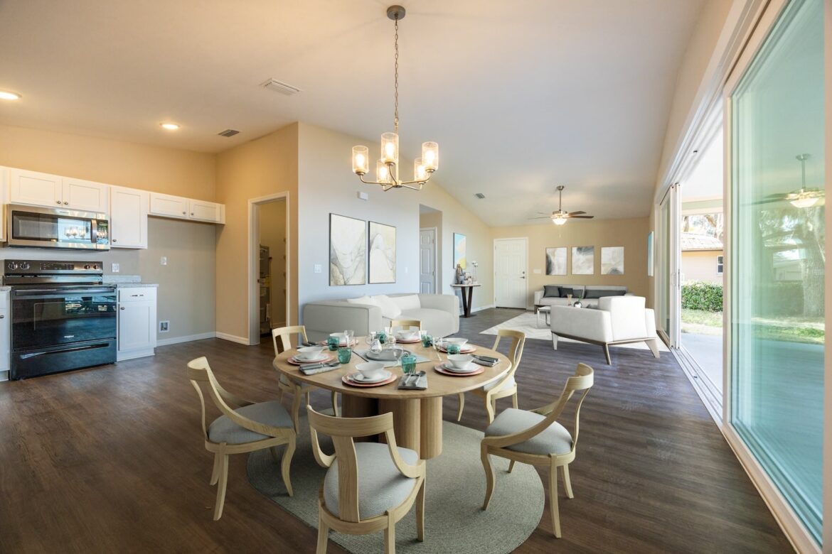 Open-concept kitchen, dining, and living area in a modern home with wood flooring, neutral walls, and contemporary furniture. Large sliding glass doors offer natural light and a view of the backyard.