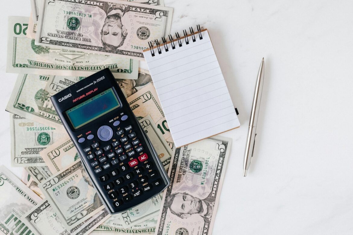 Scientific calculator, notebook and pen placed on top of scattered U.S. dollar bills, representing budgeting, financial planning or accounting tasks.