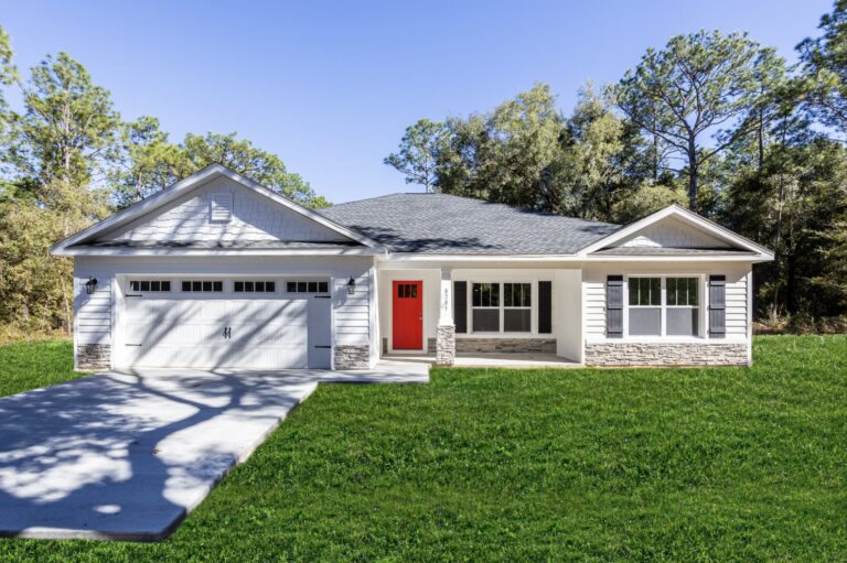 A single-story modern house with white siding, a gray roof, a bright red front door, and a two-car garage, set on a green lawn with trees in the background under a clear blue sky.