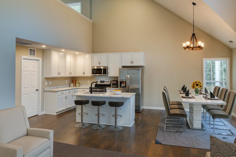 Modern open-concept kitchen and dining area with white cabinets, stainless steel appliances, an island with three bar stools, a dining table with eight chairs, and a vase of sunflowers by a large window.