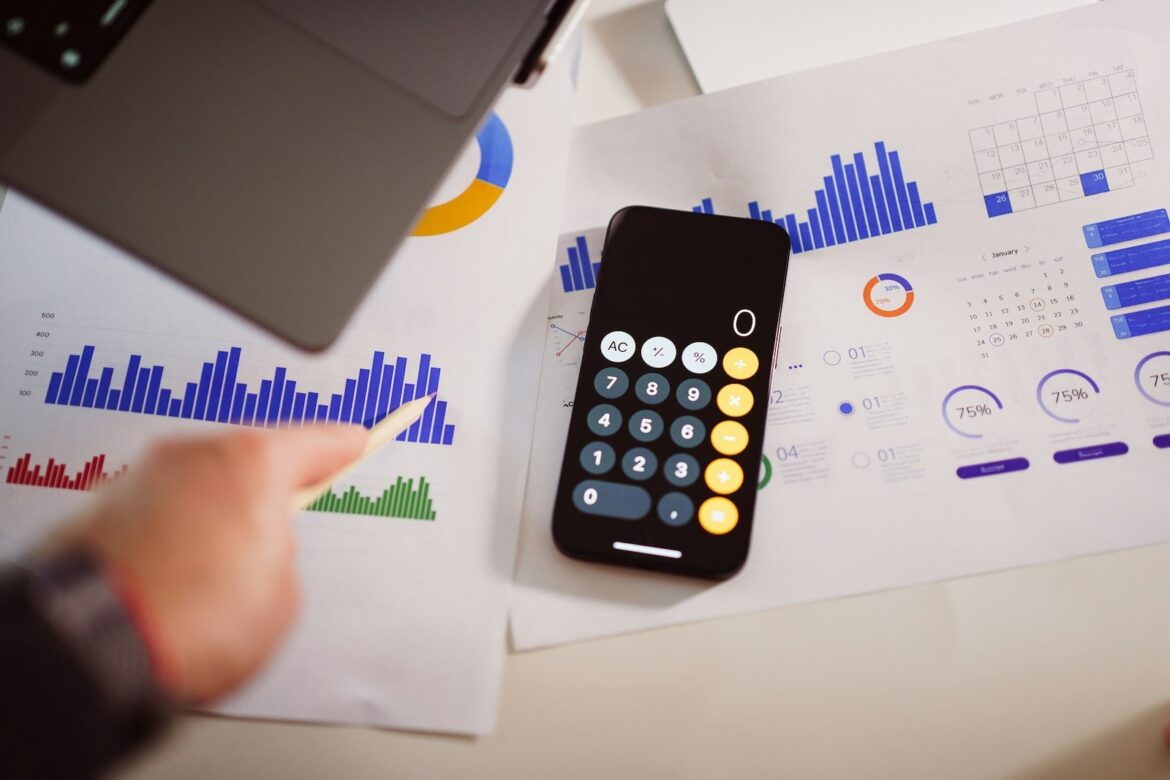 A smartphone calculator app displaying zero rests on top of printed charts and graphs with blue, green, red and orange bars. A hand holding a pencil points toward one of the bar graphs, and part of a laptop is visible in the upper corner.