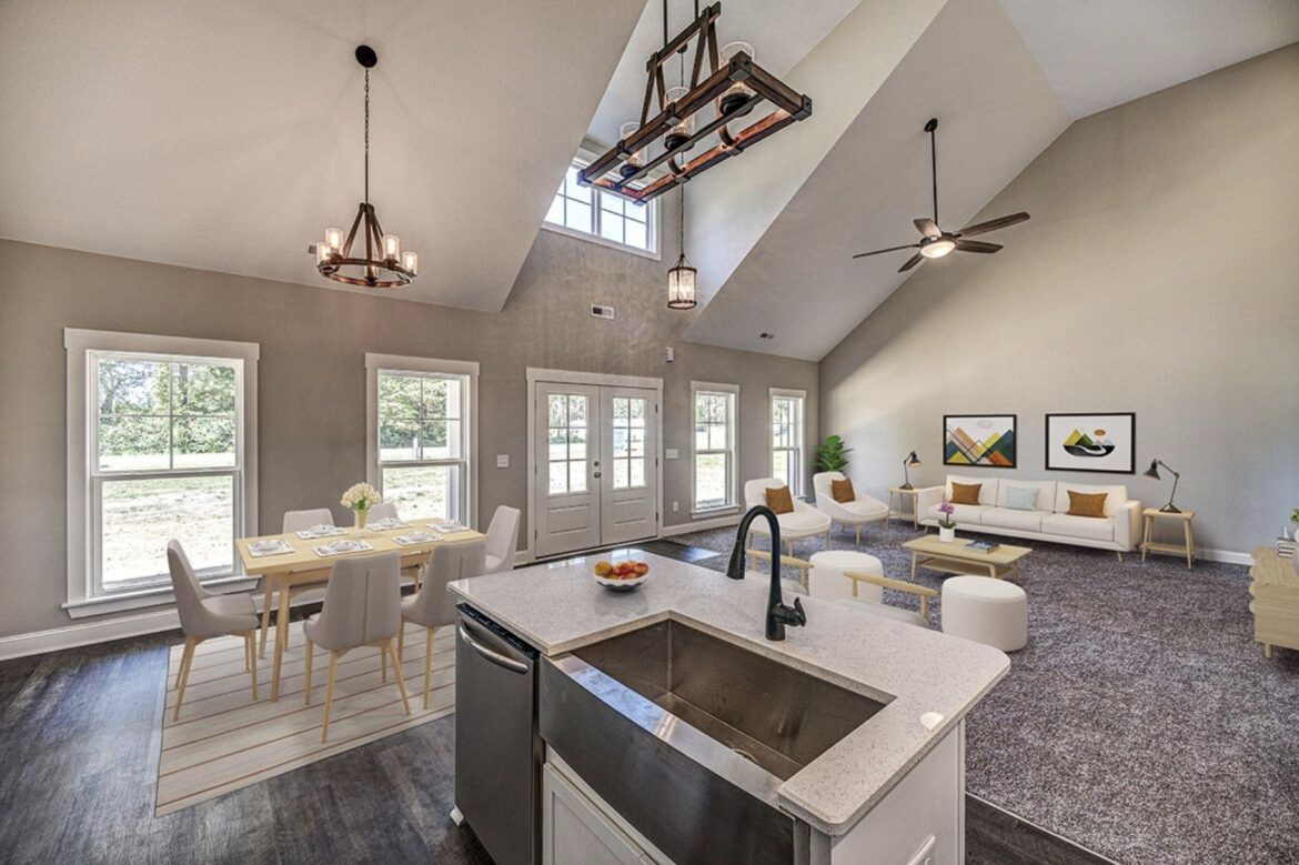 Bright open-concept living and dining area with vaulted ceilings, large windows, and modern lighting fixtures, viewed from the kitchen island with a sink in the foreground.